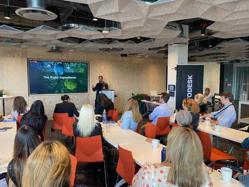 Presenter at Autodesk Partner Summit event beside a slide titled The Right Ingredients, audience seated in orange chairs