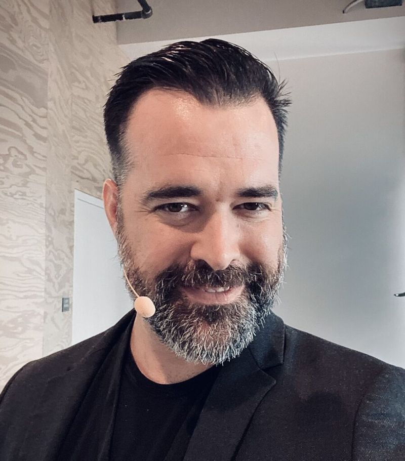 Man with beard and beige earpiece microphone wearing a black blazer, smiling at the camera